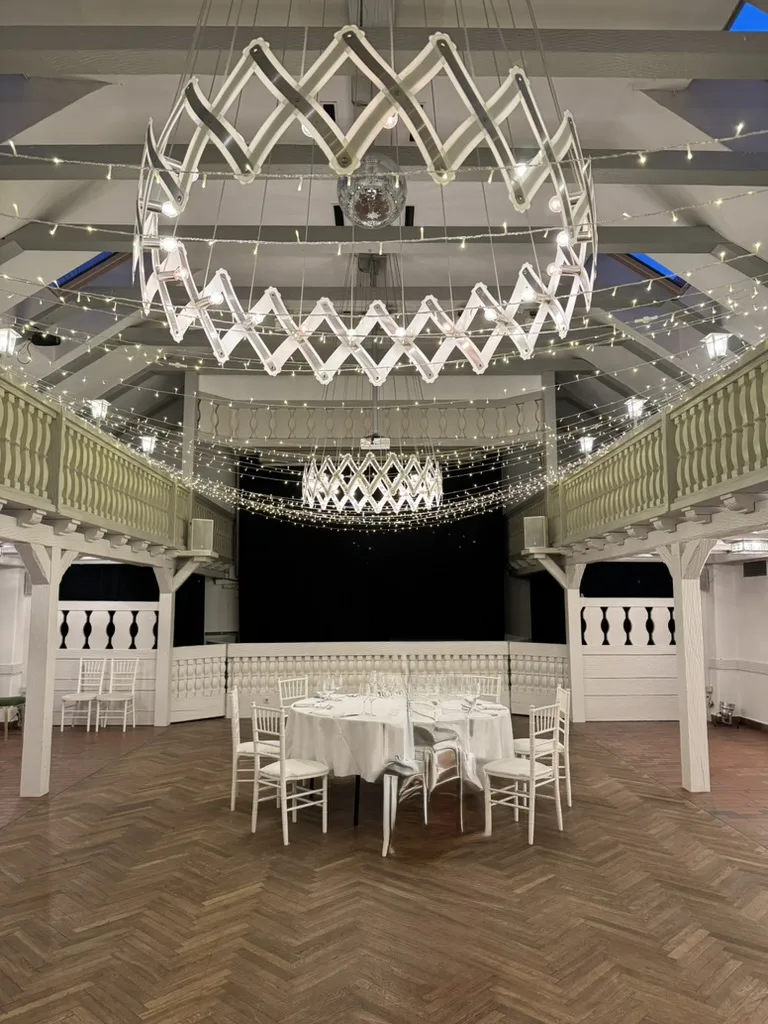 Der Drei-Kronen-Saal im Landhotel 3 Kronen in Adelsdorf mit Fischgrätparkett, umlaufender weißer Galerie, Lichterketten und einer runden Tafel vor der Bühne
